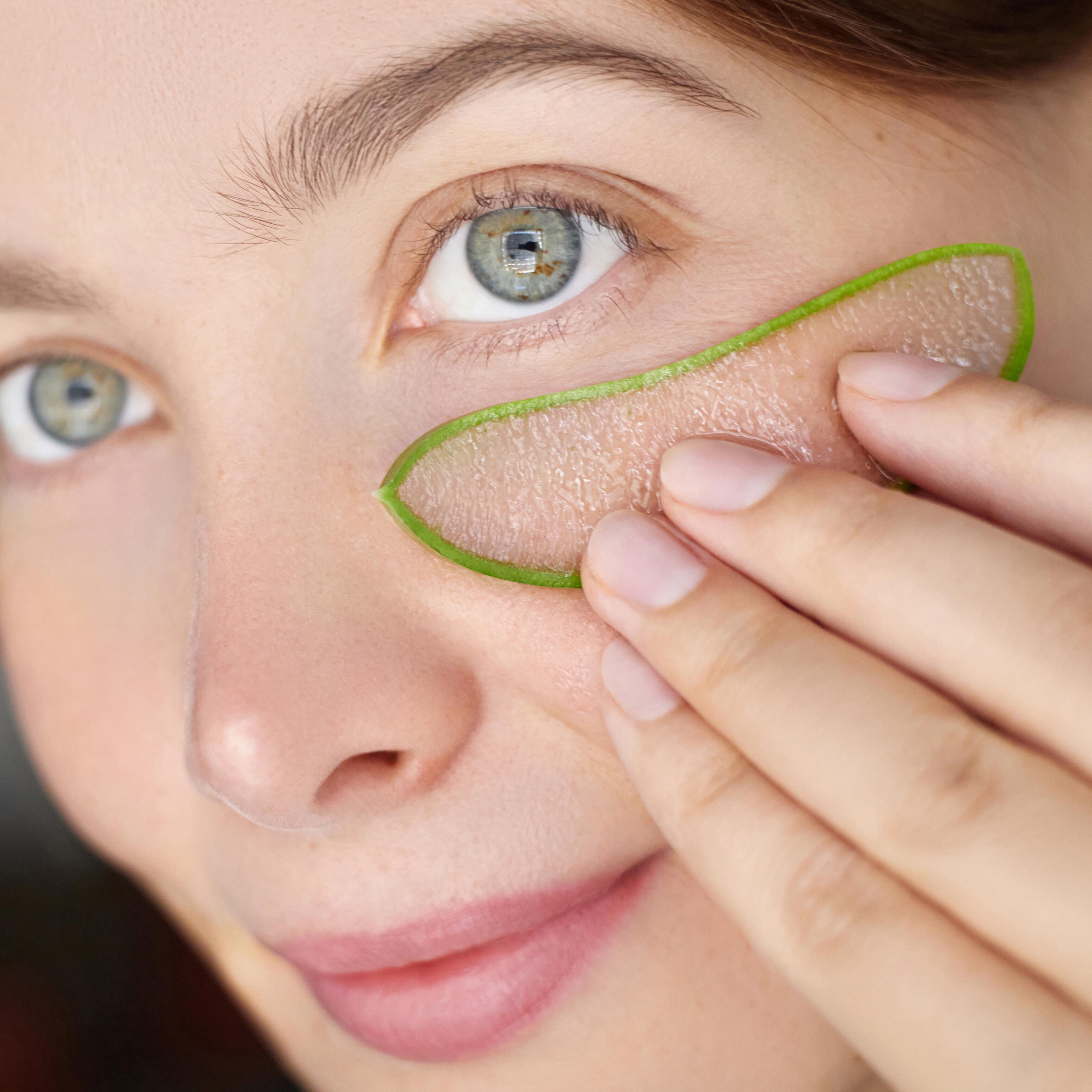 Nahaufnahme des Gesicht einer lächelnden Frau, sie hält mit einer Hand einen Teil der Aloe vera Pflanze unter ihr Auge.